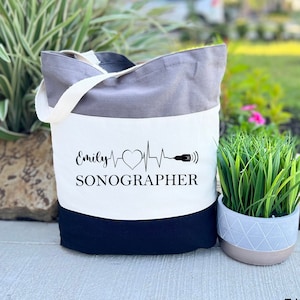 May include: A tri-color grey tote bag with a white panel featuring the text "Emily SONOGRAPHER" and a stylized heartbeat with a heart and a probe.