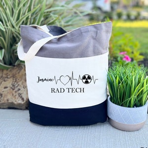 May include: A tri-color grey tote bag with a white panel featuring a black and white graphic of a heart beat with a radiation symbol and the text "Jasmine RAD TECH".