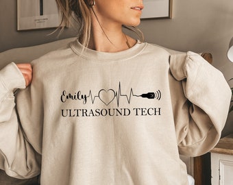May include: A sand-colored sweatshirt with a black graphic design featuring a heartbeat with a heart, a probe, and the text "Emily ULTRASOUND TECH".