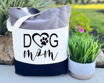 May include: A tri-color tote bag with a white panel featuring the text "DOG mom" in black with a paw print inside a heart. The bag is grey, white, and black.