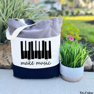 May include: A tri-color tote bag with a black and white piano keyboard graphic and the text "make music". The bag is grey, white, and black.