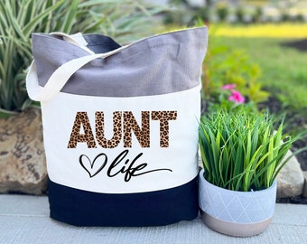 May include: A white tote bag with a black and grey striped design. The bag has a black handle and the text "AUNT life" in leopard print letters with a heart.