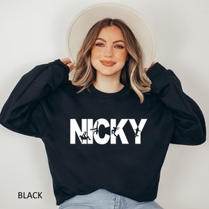 May include: Black sweatshirt with the word "NICKY" printed in white with a graphic of stick figures in a dance pose. The sweatshirt is being worn by a person with long blonde hair and a white hat.