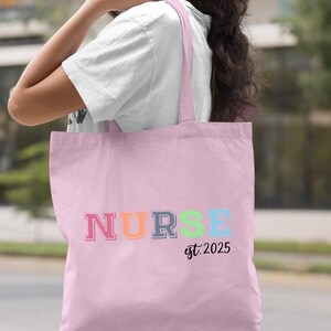 May include: Light pink tote bag with the word "NURSE" in rainbow colours and the text "est. 2025" underneath.