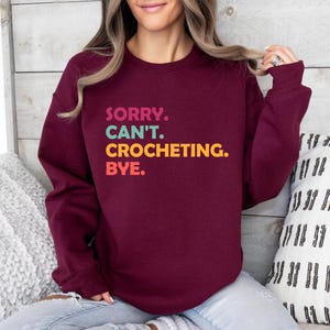 May include: A maroon sweatshirt with the text "SORRY. CAN'T. CROCHETING. BYE." in colorful letters.