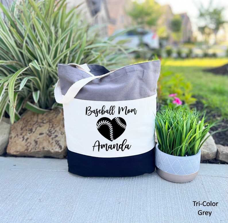 May include: A tri-color tote bag with a white panel featuring the text "Baseball Mom Amanda" and a black and white baseball heart graphic. The bag is grey, white, and black.