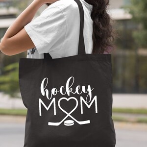 May include: Black canvas tote bag with white text that reads "hockey MOM" with a heart replacing the "O".  Two hockey sticks and a puck are below the text.
