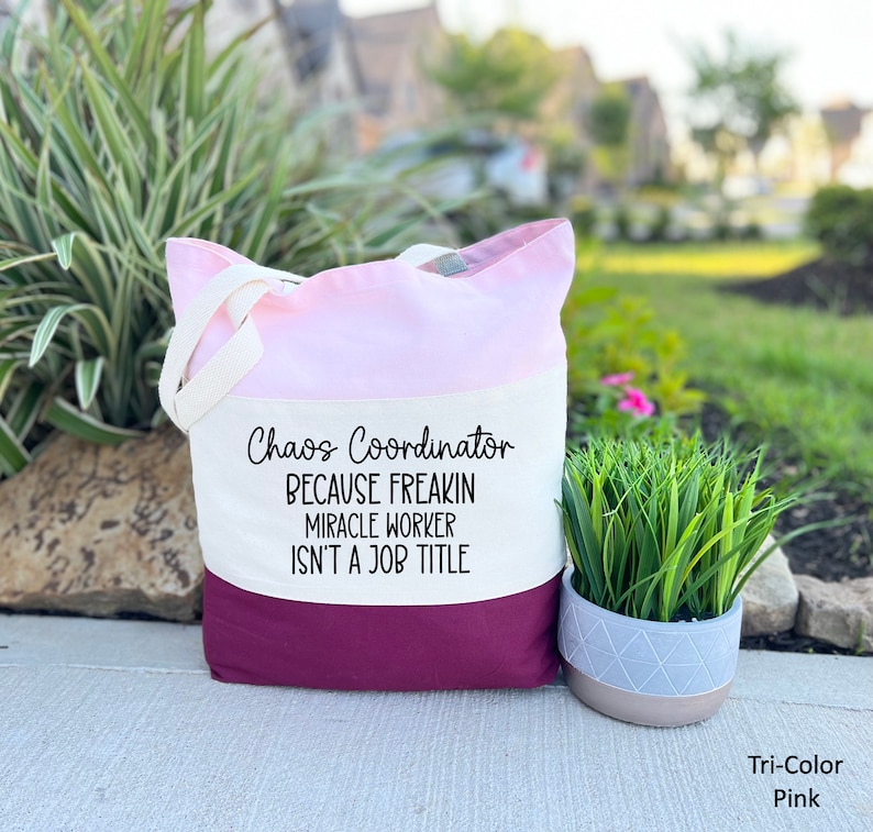 Puede incluir: Una bolsa de mano tricolor con un dise&ntilde;o rosa, blanco y burdeos. La bolsa tiene una secci&oacute;n de lona blanca con el texto "Chaos Coordinator Because Freakin Miracle Worker Isn't A Job Title".