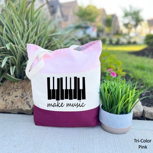 May include: A tri-color tote bag with a pink, white, and burgundy design. The bag features a black silhouette of a piano keyboard with the text "make music" underneath.