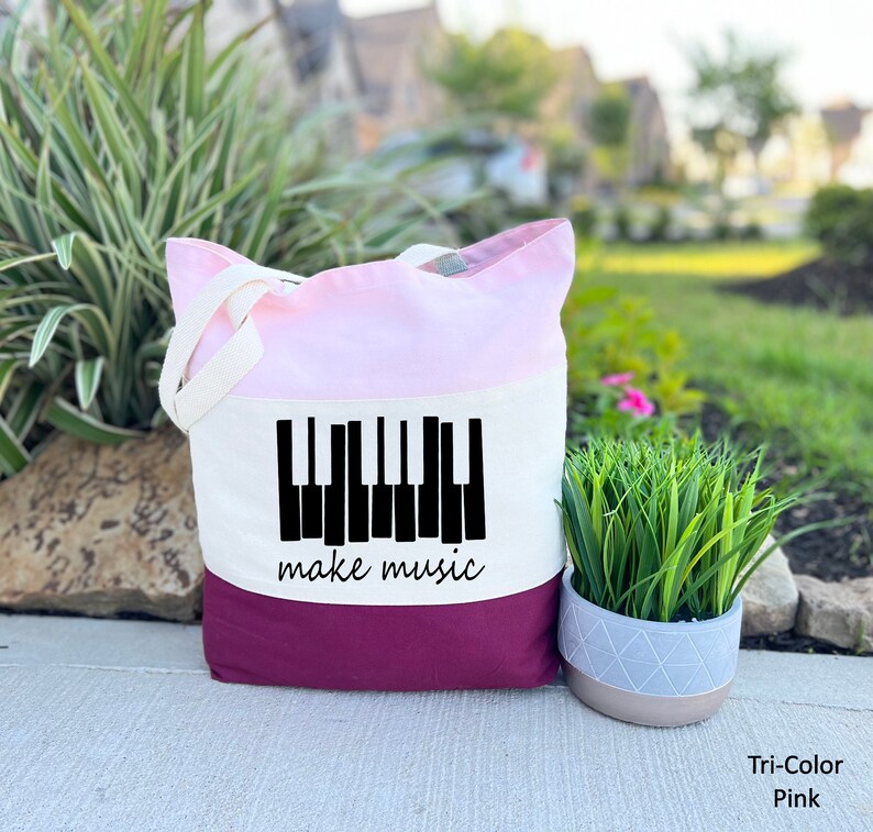 May include: A tri-color tote bag with a pink, white, and burgundy design. The bag features a black silhouette of a piano keyboard with the text "make music" underneath.