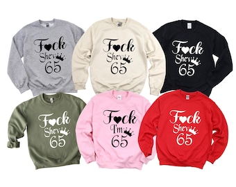 Puede incluir: Seis sudaderas de cuello redondo en varios colores: gris, beige, negro, verde, rosa y rojo. Cada sudadera presenta el texto "Fock She's 65" en fuente negra con un diseño de corazón y corona.