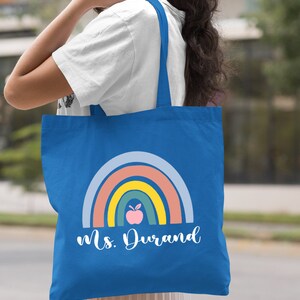 Puede incluir: Una bolsa de tela azul real con un dise&ntilde;o de arco&iacute;ris y una manzana. El texto "Ms. Durand" est&aacute; impreso en la bolsa.