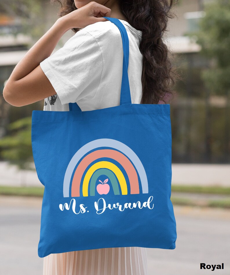 Puede incluir: Una bolsa de tela azul real con un dise&ntilde;o de arco&iacute;ris y una manzana. El texto "Ms. Durand" est&aacute; impreso en la bolsa.