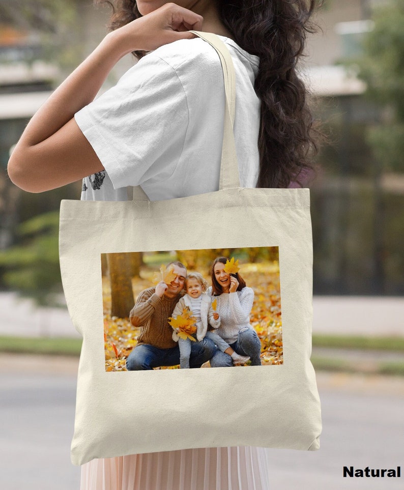 May include: A white canvas tote bag with a photo of a family of three in a park setting. The family is smiling and holding leaves in front of their faces. The background is a blurred image of trees and leaves.