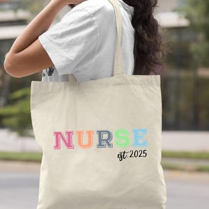 May include: A natural canvas tote bag with the word "NURSE" in rainbow colours and the text "est. 2025" in black.