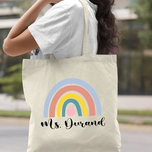 Puede incluir: Una bolsa de lona natural con un dise&ntilde;o de arco&iacute;ris y el texto "Ms. Durand". El arco&iacute;ris tiene una manzana rosa en el centro.