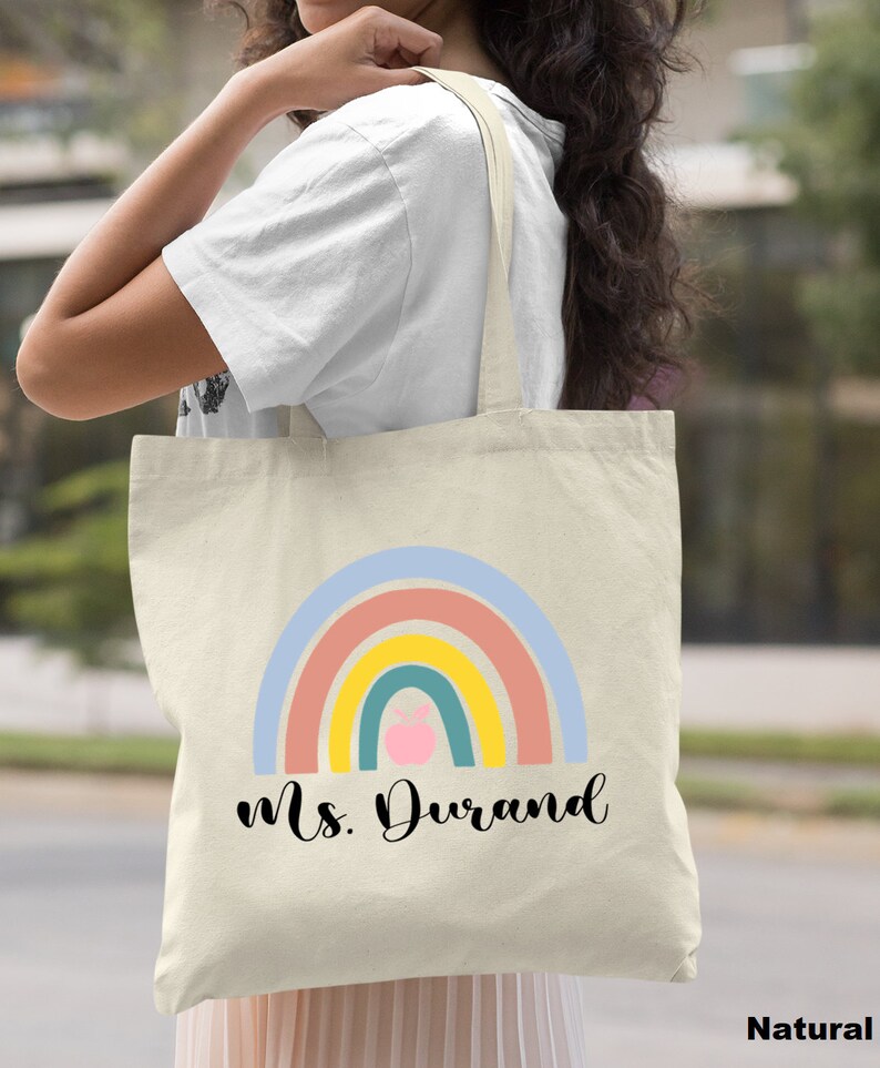 Puede incluir: Una bolsa de lona natural con un dise&ntilde;o de arco&iacute;ris y el texto "Ms. Durand". El arco&iacute;ris tiene una manzana rosa en el centro.
