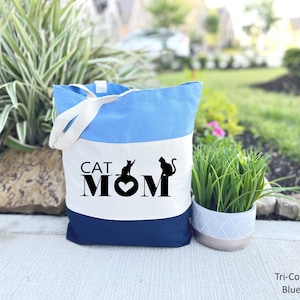 Peut inclure: Un sac fourre-tout tricolore avec un design bleu, blanc et bleu marine. Le sac porte le texte "CAT MOM" avec deux silhouettes de chats et un c&oelig;ur au centre.