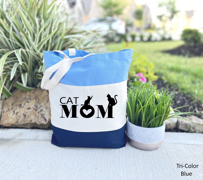 Peut inclure: Un sac fourre-tout tricolore avec un design bleu, blanc et bleu marine. Le sac porte le texte "CAT MOM" avec deux silhouettes de chats et un c&oelig;ur au centre.