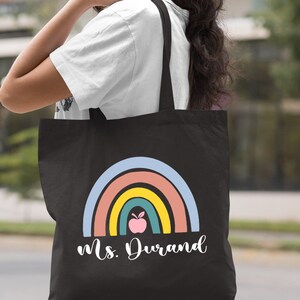 Puede incluir: Bolsa de lona negra con un dise&ntilde;o de arco&iacute;ris y el texto "Ms. Durand". El arco&iacute;ris tiene una manzana rosa en el centro.