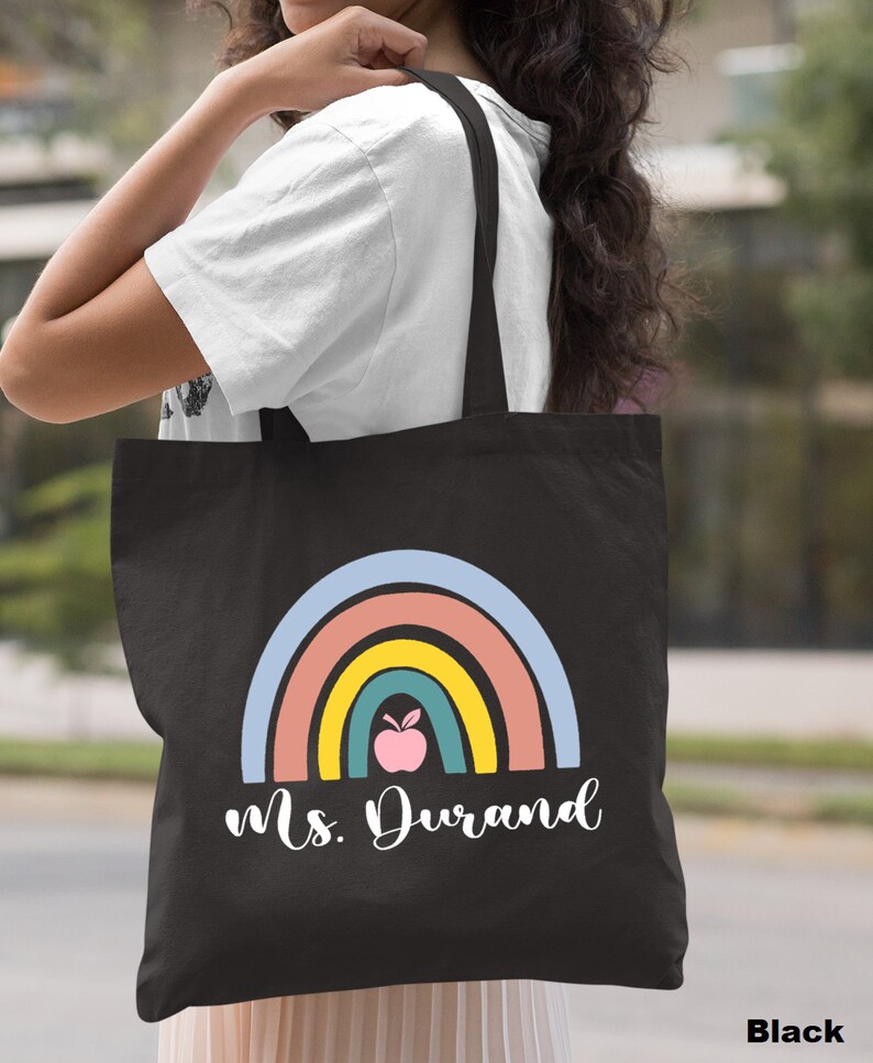 Puede incluir: Bolsa de lona negra con un dise&ntilde;o de arco&iacute;ris y el texto "Ms. Durand". El arco&iacute;ris tiene una manzana rosa en el centro.