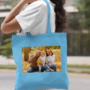 May include: A light blue tote bag with a photo of a family of three in a fall setting. The family is smiling and holding leaves in front of their faces. The photo is printed on the front of the bag.