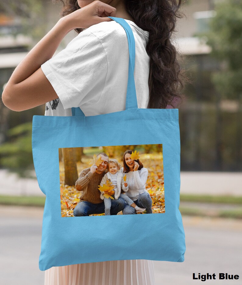 May include: A light blue tote bag with a photo of a family of three in a fall setting. The family is smiling and holding leaves in front of their faces. The photo is printed on the front of the bag.