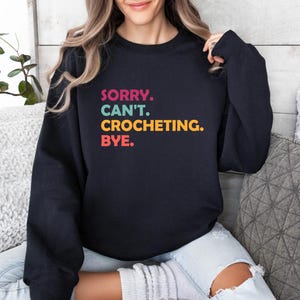 May include: A black sweatshirt with the text "SORRY. CAN'T. CROCHETING. BYE." in colorful letters.