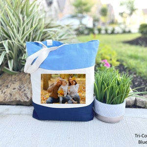 May include: A blue, white, and navy blue canvas tote bag with a photo of a family of three in a fall setting. The photo shows a man, a woman, and a child smiling and holding leaves.