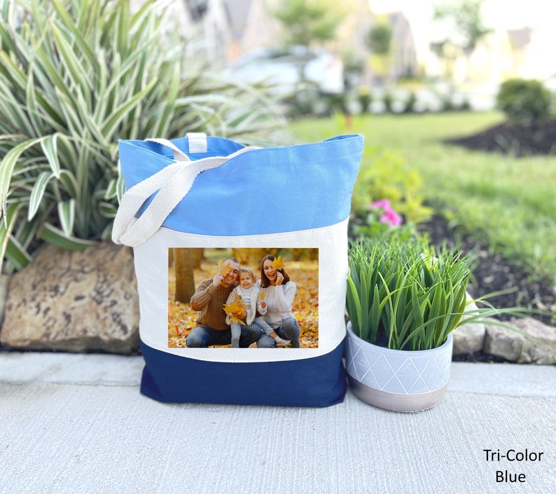 May include: A blue, white, and navy blue canvas tote bag with a photo of a family of three in a fall setting. The photo shows a man, a woman, and a child smiling and holding leaves.
