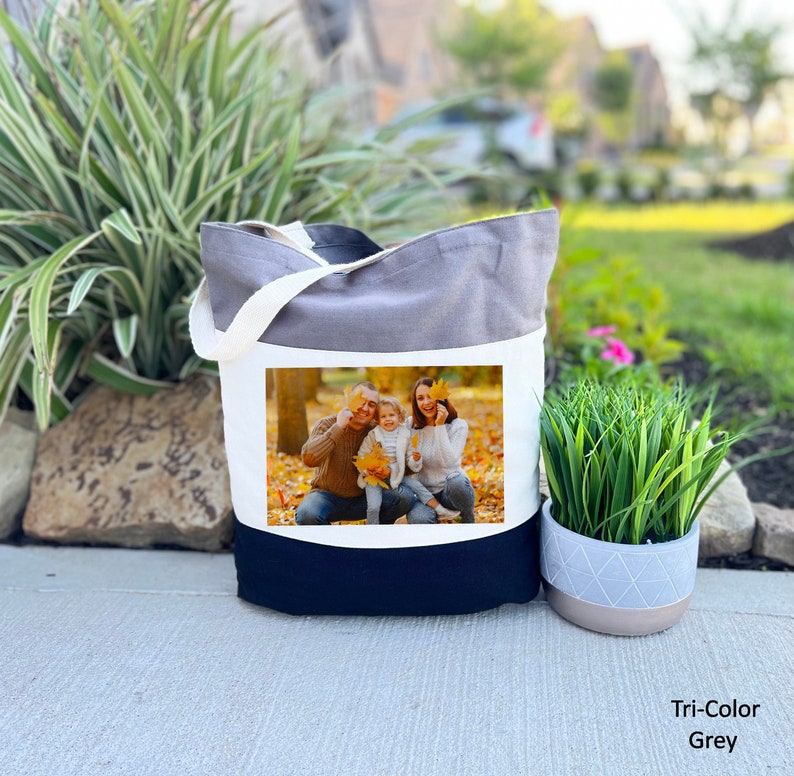May include: A grey, white, and navy blue tote bag with a photo of a family of three in a fall setting. The photo shows a man, a woman, and a child standing in a park with yellow leaves on the ground.