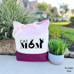 Peut inclure: Un sac fourre-tout tricolore avec un design rose, blanc et bordeaux. Le sac pr&eacute;sente le texte "CAT MOM" avec deux silhouettes de chats noirs et un c&oelig;ur.