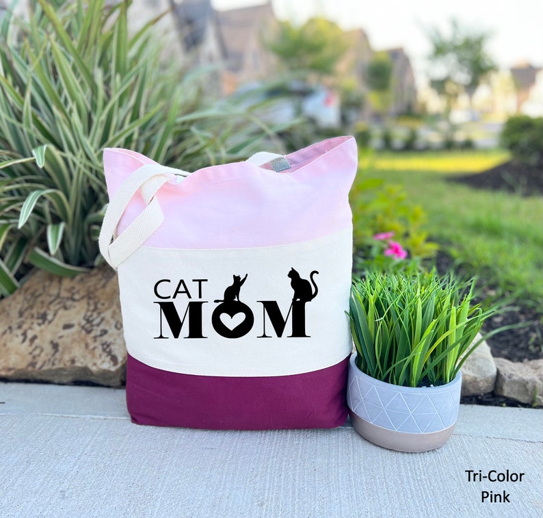 Peut inclure: Un sac fourre-tout tricolore avec un design rose, blanc et bordeaux. Le sac pr&eacute;sente le texte "CAT MOM" avec deux silhouettes de chats noirs et un c&oelig;ur.