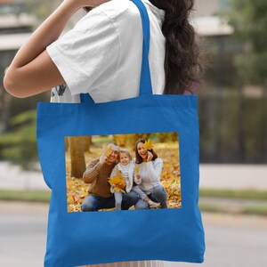 May include: A royal blue tote bag with a photo of a family of three in a park setting. The family is smiling and holding autumn leaves. The bag has a white strap.