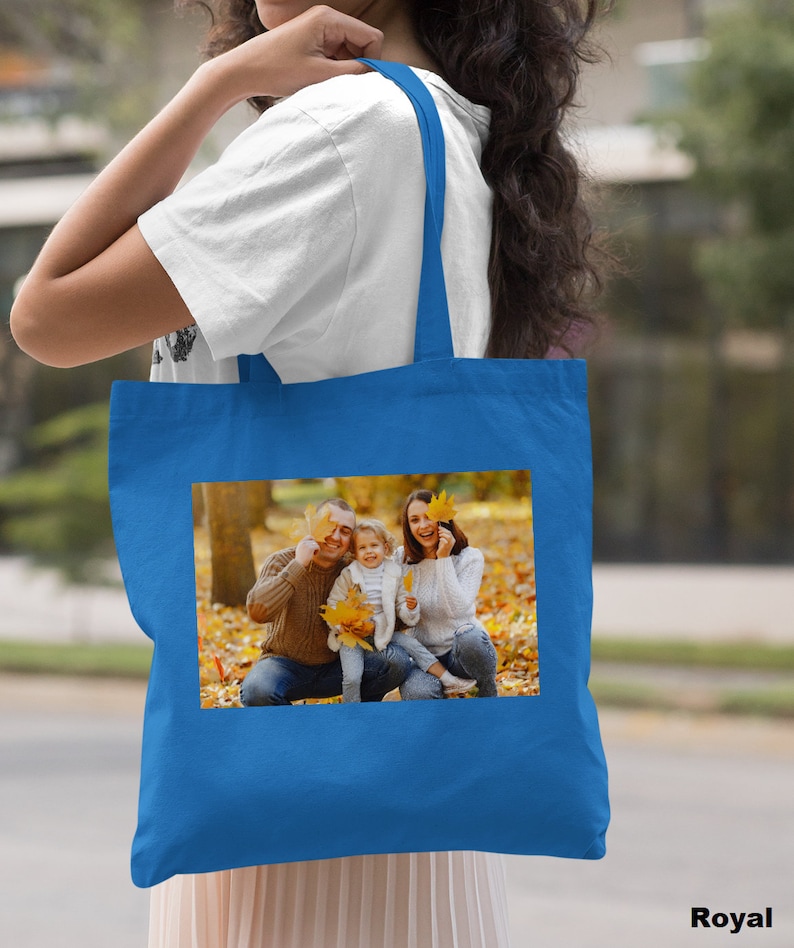 May include: A royal blue tote bag with a photo of a family of three in a park setting. The family is smiling and holding autumn leaves. The bag has a white strap.