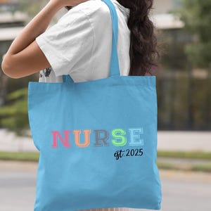 May include: Light blue tote bag with the word "NURSE" in rainbow colours and the text "est. 2025" in black.