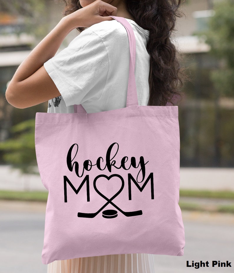 May include: Light pink canvas tote bag with black text that reads "hockey MOM" with a heart in place of the "O". Two black hockey sticks are crossed below the text with a black hockey puck in the center.