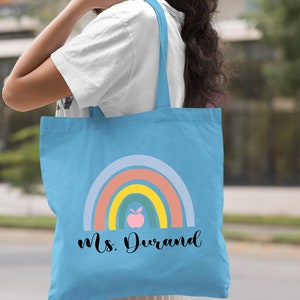 Puede incluir: Bolsa de tela azul claro con un dise&ntilde;o de arco&iacute;ris y el texto "Ms. Durand" en negro. El arco&iacute;ris tiene un esquema de color rosa, naranja, amarillo, verde y azul. Hay una manzana rosa con una hoja verde en el centro del arco&iacute;ris.