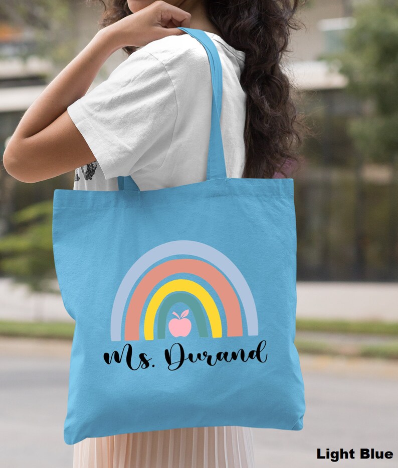 Puede incluir: Bolsa de tela azul claro con un dise&ntilde;o de arco&iacute;ris y el texto "Ms. Durand" en negro. El arco&iacute;ris tiene un esquema de color rosa, naranja, amarillo, verde y azul. Hay una manzana rosa con una hoja verde en el centro del arco&iacute;ris.