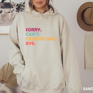 May include: A light beige hoodie with the text "SORRY. CAN'T. CROCHETING. BYE." printed in rainbow colors.