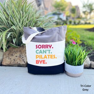 May include: A gray, white, and black canvas tote bag with the text "SORRY. CAN'T. PILATES. BYE." printed in colorful letters. The bag has a white canvas strap.