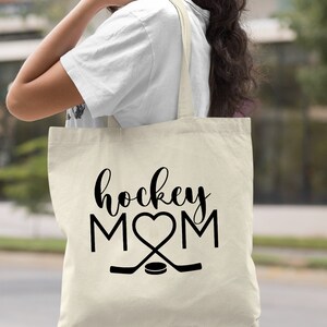 May include: A natural canvas tote bag with black text that reads "hockey MOM" with a heart in place of the "O". Two hockey sticks are crossed below the text with a puck in the center.