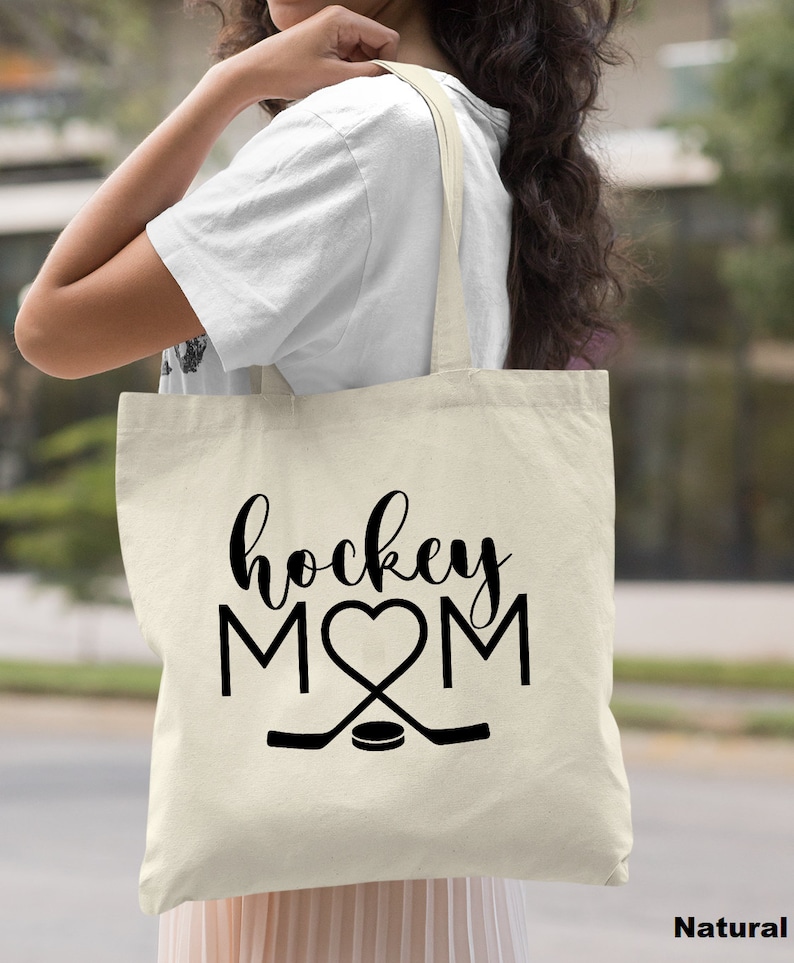 May include: A natural canvas tote bag with black text that reads "hockey MOM" with a heart in place of the "O". Two hockey sticks are crossed below the text with a puck in the center.