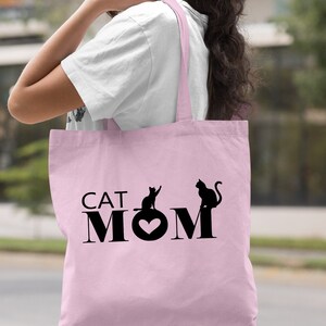 Peut inclure: Sac fourre-tout rose p&acirc;le avec du texte noir qui dit "CAT MOM" avec un c&oelig;ur au milieu du "O". Deux silhouettes de chats noirs sont de chaque c&ocirc;t&eacute; du texte.