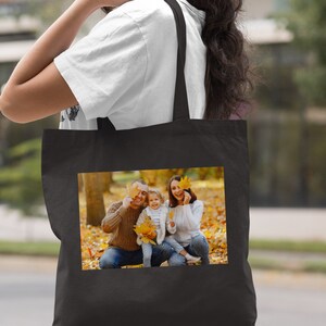 May include: Black tote bag with a photo of a family of three in a park setting. The family is smiling and holding autumn leaves. The bag has a black handle.