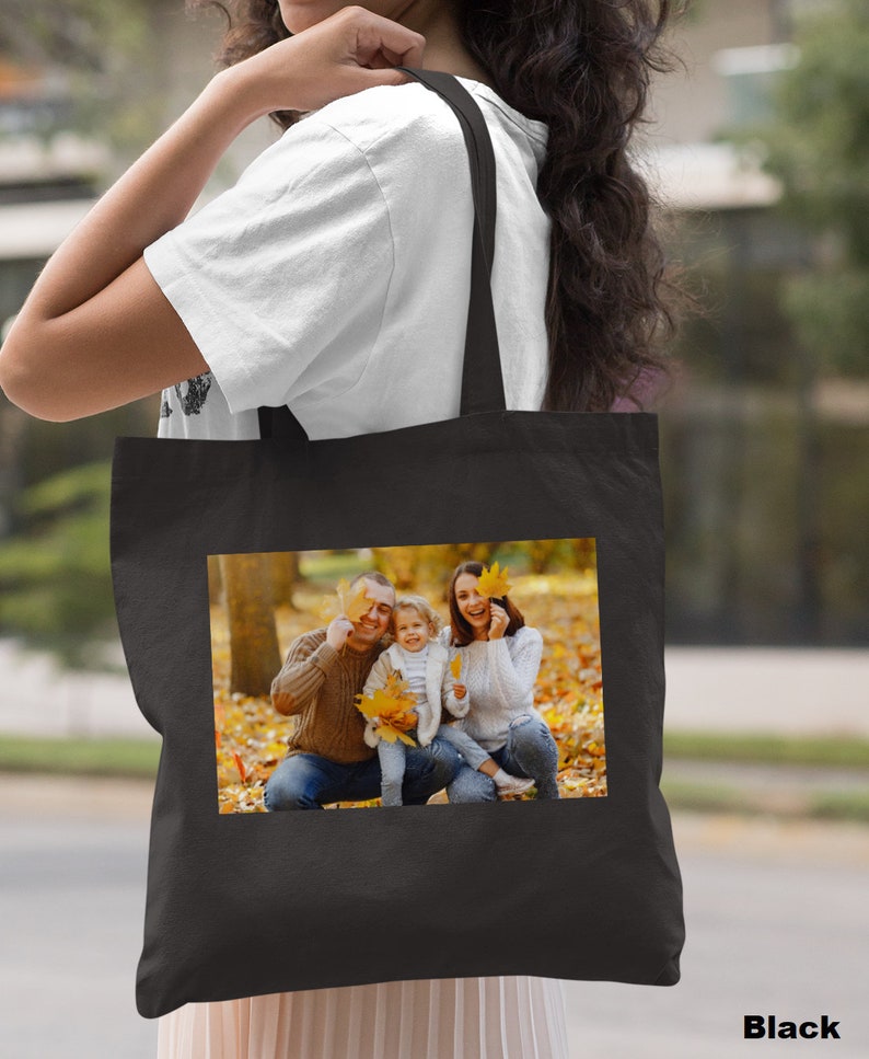 May include: Black tote bag with a photo of a family of three in a park setting. The family is smiling and holding autumn leaves. The bag has a black handle.
