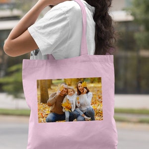 May include: A light pink tote bag with a photo of a family of three in a park setting. The family is smiling and holding leaves in front of their faces. The bag has a long handle and is made of canvas.