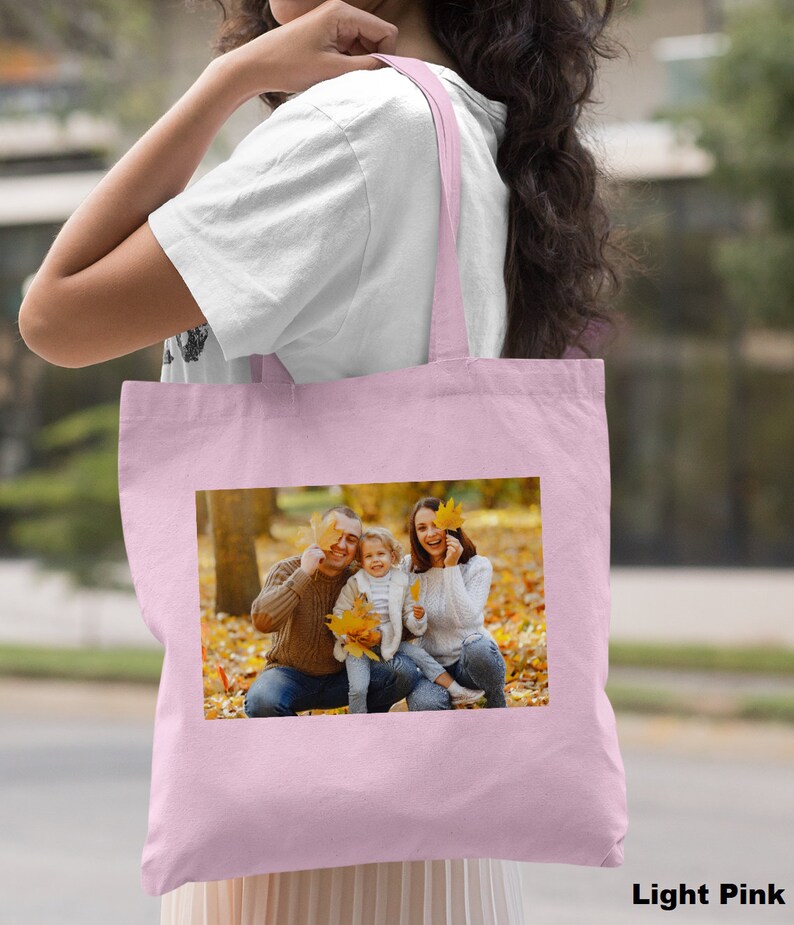 May include: A light pink tote bag with a photo of a family of three in a park setting. The family is smiling and holding leaves in front of their faces. The bag has a long handle and is made of canvas.