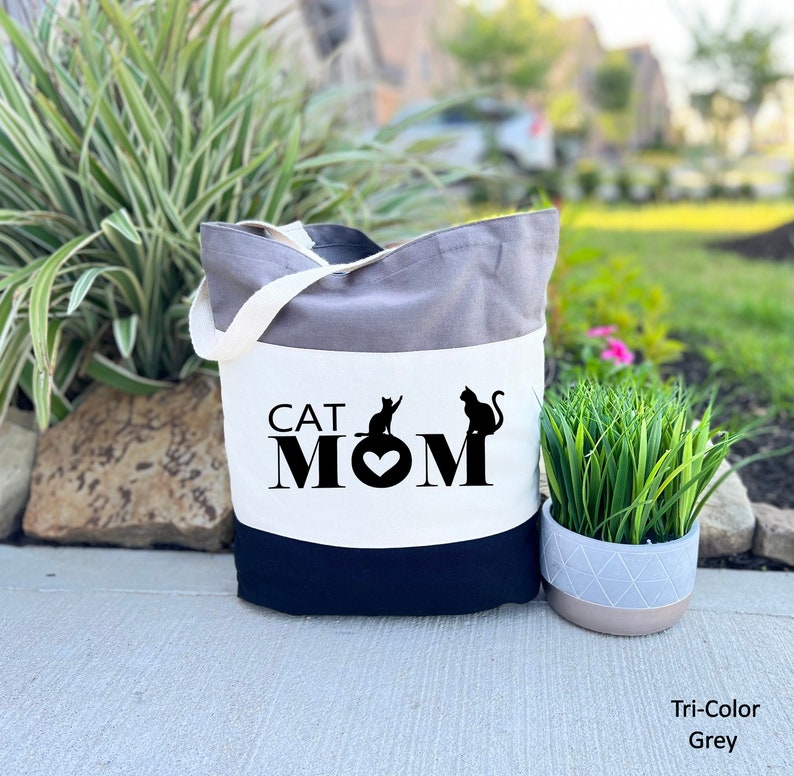Peut inclure: Un sac fourre-tout gris tricolore avec un panneau blanc sur lequel est inscrit le texte "CAT MOM" avec deux silhouettes de chats noirs et un c&oelig;ur.