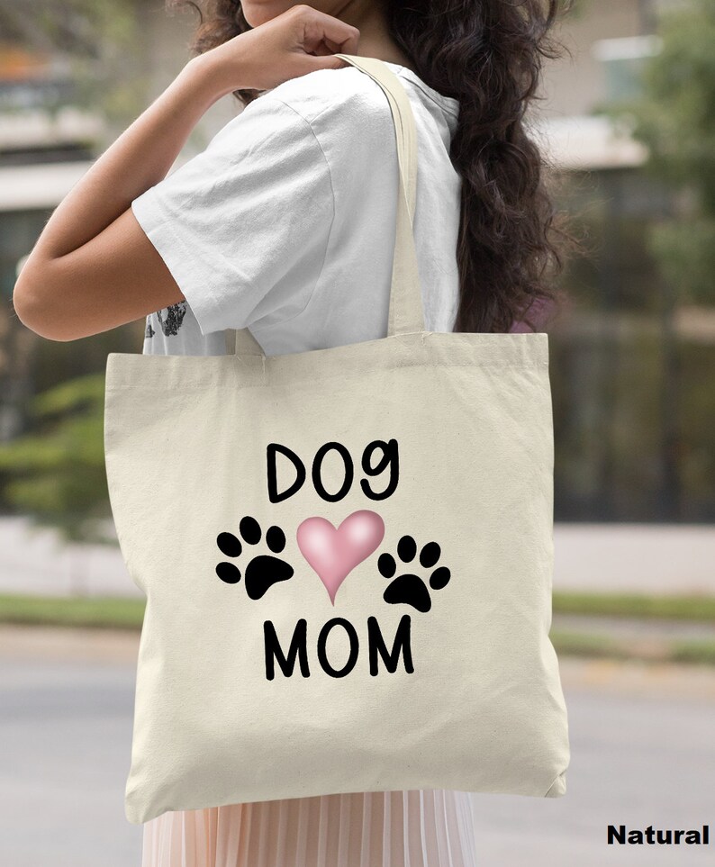 May include: A natural canvas tote bag with black text that reads "Dog Mom" and features a pink heart and two black paw prints.